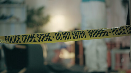 Close-up of crime scene tape, with forensic experts examining the room and collecting evidence