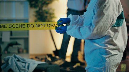 A forensic expert is marking the murder scene with warning tape, and police detectives are...