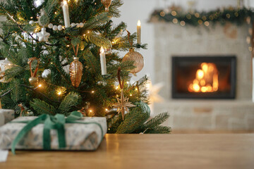 Merry Christmas! Stylish christmas decorated tree, golden lights, gift on table and burning fireplace. Atmospheric christmas eve at fireplace, space for text - obrazy, fototapety, plakaty