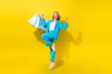 Full length photo of ecstatic woman wear blue stylish suit hold shopping bags jumping celebrate...