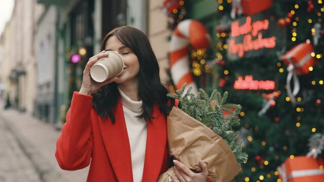 Caucasian woman in festive mood walking in the winter decorated street, drinking coffee or tea and a bouquet of christmas tree. Brunette woman walking in the city center. The winter holidays.