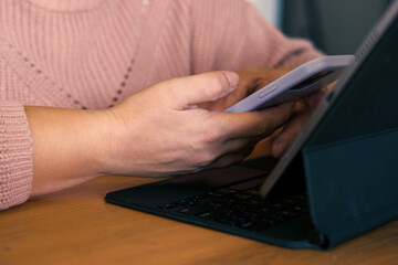 Female using phone looking at laptop or digital tablet device. Unrecognizable woman sitting at table in a cafe and use online banking with smartphone. Easy pay. App for sale in shop. Hands close up