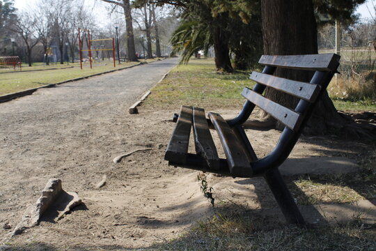 Banco de parque de hierro y madera debajo del &aacute;rbol con sol y sombra, para sentarse y relajarse, dando sensaci&oacute;n de calma, se encuentra solitario con fondo del camino de ripio y los &aacute;rboles