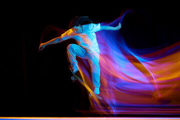 Man skateboarder performing freestyle tricks in motion isolated black studio background in mixed neon flashes of light. Concept of youth culture.