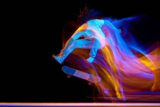 Full-length portrait of man dressed modern stylishly outfit skateboarding with neon flashes of light isolated black background.