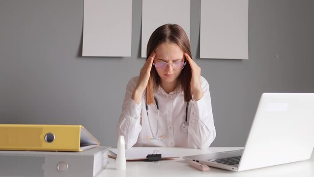 Tired woman doctor with stethoscope wearing white lab coat sitting in her office at table with laptop has many patients working long hours in front of computer suffering headache.