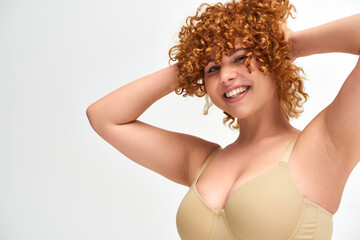 Obraz premium cheerful young woman with curvy breasts touching red wavy hair on white backdrop, plus size beauty