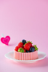 Tartlet with strawberries, raspberries, blueberries, basket of pink chocolate and heart on a pink background.