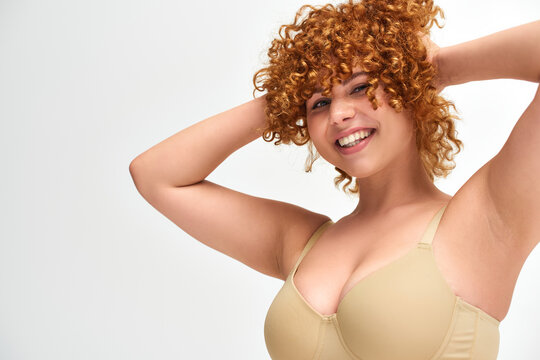 cheerful young woman with curvy breasts touching red wavy hair on white backdrop, plus size beauty