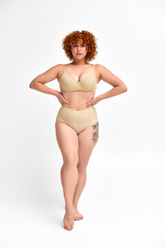 full length of plus size tattooed model in beige lingerie with hands on hips on white backdrop