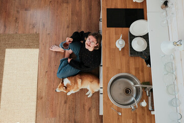 A tall blonde man sitting on a floor in the kitchen and drinks tea in the company of his dog