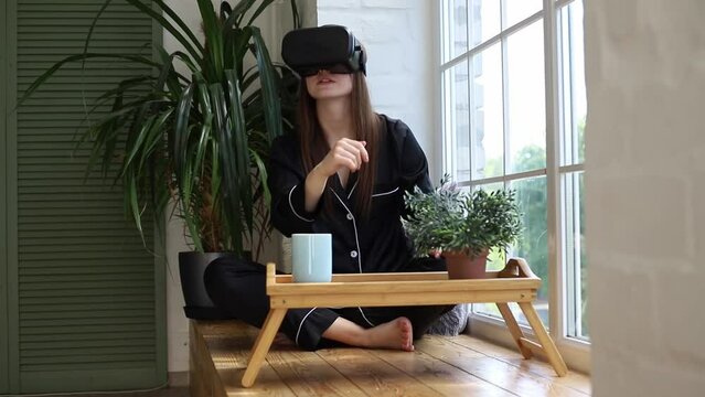 Young woman sitting at breakfast on the windowsill wearing virtual glasses, watching a simulation of a game or a fantasy world. The concept of leaving real life for virtual metaverse life.