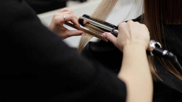 Unrecognisable professional hairdresser ironing female client hair with hair straightener and comb, making stylish hairstyle in beauty salon. Keratin, lamination,hair care, hair salon and hairstyling.