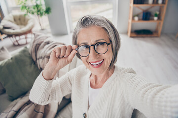Photo of cheerful positive elderly lady wear white cardigan spectacles tacking selfie smiling indoors apartment room