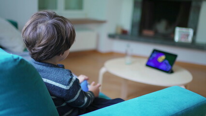 Child watching cartoon at home seated on sofa couch, small boy absorbed by entertainment content on tablet device screen