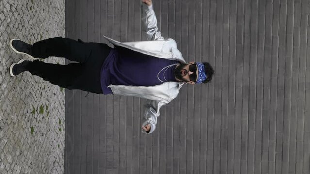 A vertical shot captures a man, dressed in urban style, performing freestyle hip hop dance outdoors, showcasing unique moves with a shiny jacket, sunglasses, and headband