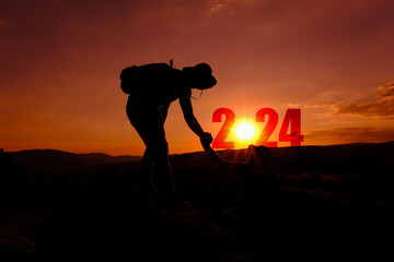 New year 2024 and new victories in business. Silhouette of man extending his hand to his friend on top of the mountain and the background shows the beginning of the New Year 2024.