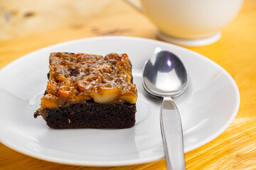 High angle view of a piece of delicious homemade toffee cake and metal spoon in white ceramic dish with a cup of coffee on wooden table.