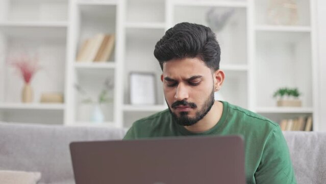 Casually dressed young guy scratching bearded chin while looking on laptop screen with pensive facial expression. Indian man sitting on couch in doubt during online work at home.
