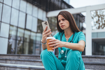Shot of a young doctor looking distressed. Stressed modern medical practitioner woman in scrubs with stethoscope, medical mask and cup of coffee outside near clinic.