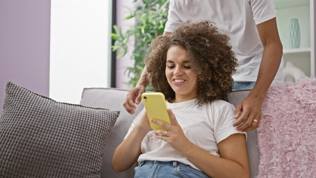 Beautiful couple enjoy laughing while sitting on the sofa at home, confidently texting light-hearted messages on their smartphone.