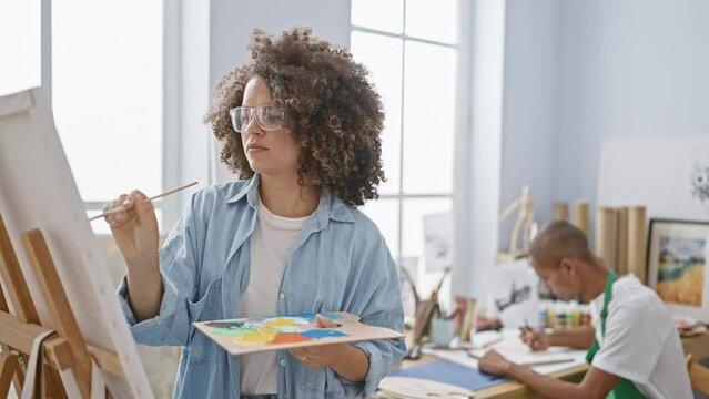 Young man and beautiful woman artists immersed in a serious drawing lesson together, standing in a relaxed art studio wielding paintbrushes over a canvas.