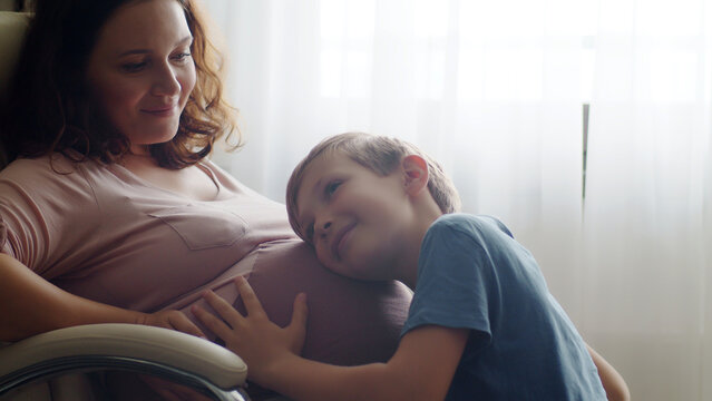 Child hugs her mother's pregnant belly. Happy son strokes the big belly his pregnant mom near window at home. Happy family life in an expectation a newborn. Pregnancy and maternity.