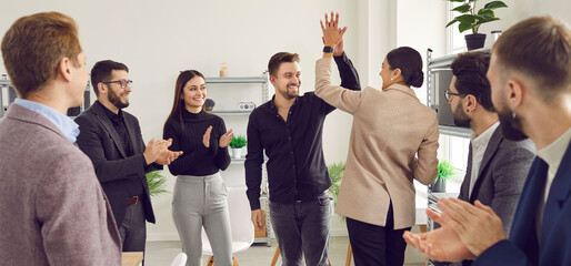 Fototapeta na wymiar Two young men doing high five while their cheerful coworkers applauding them at meeting. Colleagues celebrating achievement, victory, successful deal in conference room in office