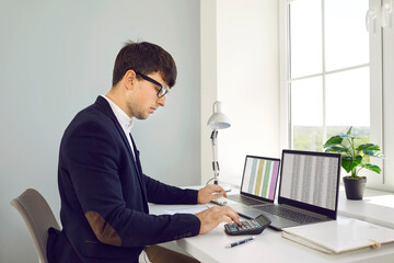 Young professional Caucasian man accountant wearing glasses uses calculator to prepare company's...