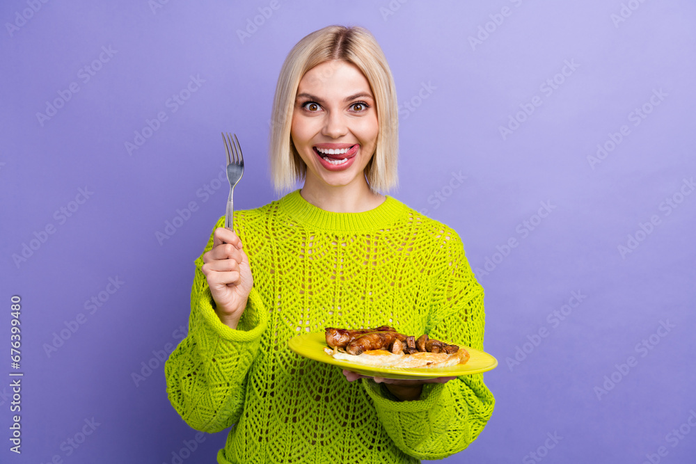 Poster Photo of hungry impressed woman dressed knitted sweater holding breakfast plate licking lips isolated violet color background