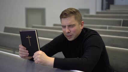 A young man prays holding a bible in his hands in a church. A Protestant man sits on a church bench and sincerely prays.