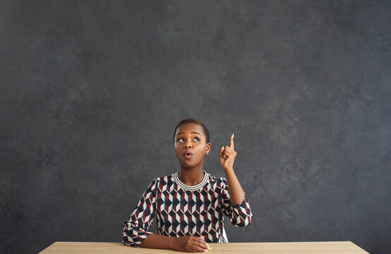 Funny dark skinned woman thinking about something sitting at the table with raised finger on gray background. Woman looking up thinking about an idea or thought that came to her mind. Place for text.