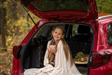 A beautiful girl with long hair sits in the trunk of a car. A girl drinks tea and warms herself in a blanket in the fall.