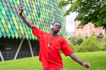 Young black man, happy dancing and listening to music with white headphones and a mobile phone in a park.