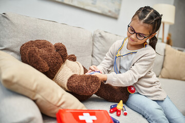 happy kid in casual wear and eyeglasses playing doctor with teddy bear on sofa in living room - obrazy, fototapety, plakaty