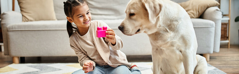 happy girl playing with colorful toy blocks near labrador in living room, building tower game banner