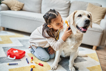 cute girl in casual attire playing doctor with labrador in modern living room, toy first aid kit
