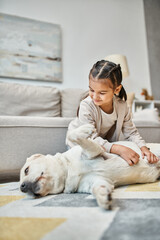 positive girl in casual attire smiling and stroking dog in modern living room, kid and labrador