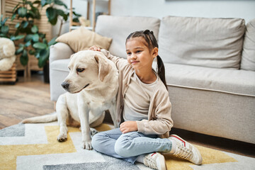cute girl smiling and sitting near sofa while stroking labrador in modern living room, kid and dog