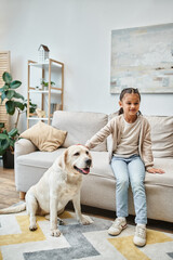 cute girl smiling and sitting on sofa while stroking labrador in modern living room, kid and dog