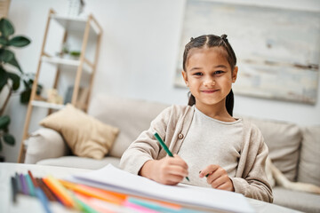 portrait of happy elementary age girl drawing with color pencil and smiling in modern apartment