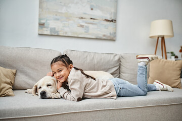 happy girl smiling and lying on sofa with cute labrador in modern living room, pet and kid