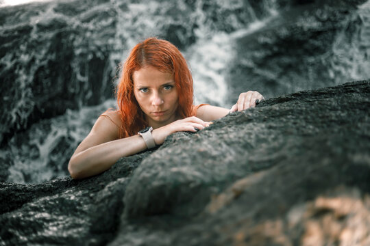 A beautiful girl sits on the stones in the jungle. Girl with red hair. Waterfall in the background.