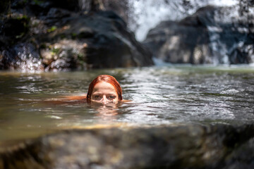 A beautiful girl swims in the lake. Waterfall in the background. Portrait of a red-haired girl.