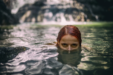 A beautiful girl swims in the lake. Waterfall in the background. Portrait of a red-haired girl.