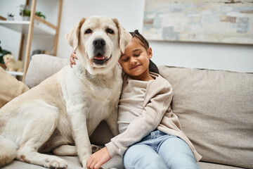 happy girl sitting on comfortable couch with cute labrador in modern living room, animal companion