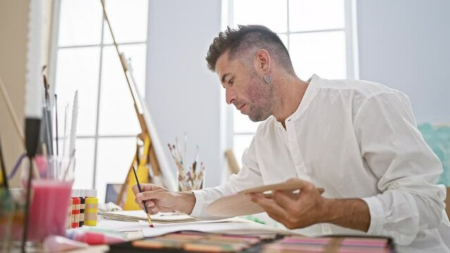Young, handsome hispanic man ardently drawing on paper, deeply engrossed and concentrated in his art at a buzzing studio