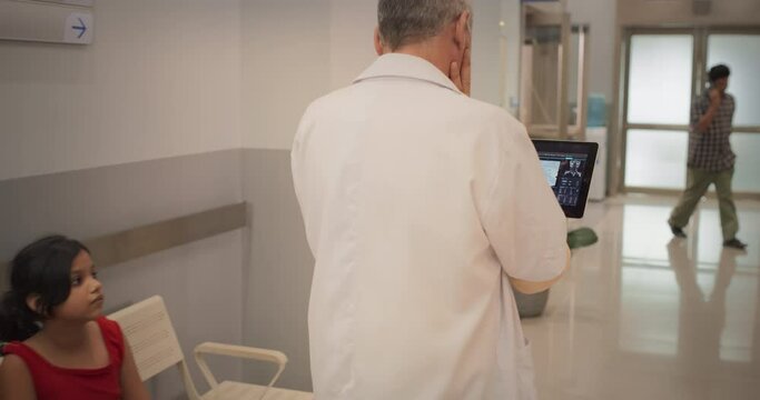 Backview Tracking Shot of Indian Doctor Walking in Hospital Corridor, Using a Digital Tablet. Slow Motion of Senior Male Surgeon Checking Brain MRI Images Before Surgery, Revisiting his Notes