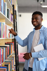 A happy and smiling young student in a university library, surrounded by books and bookshelves,...