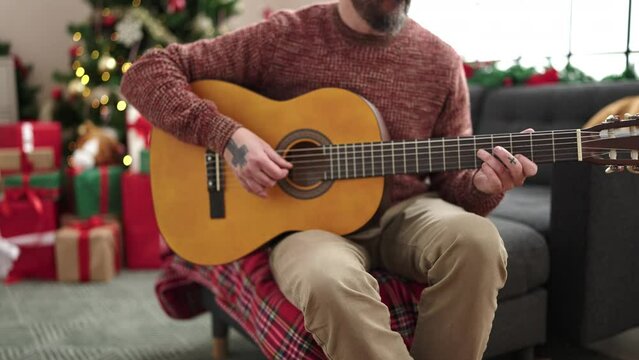Young bald man playing classical guitar sitting on sofa by christmas tree at home